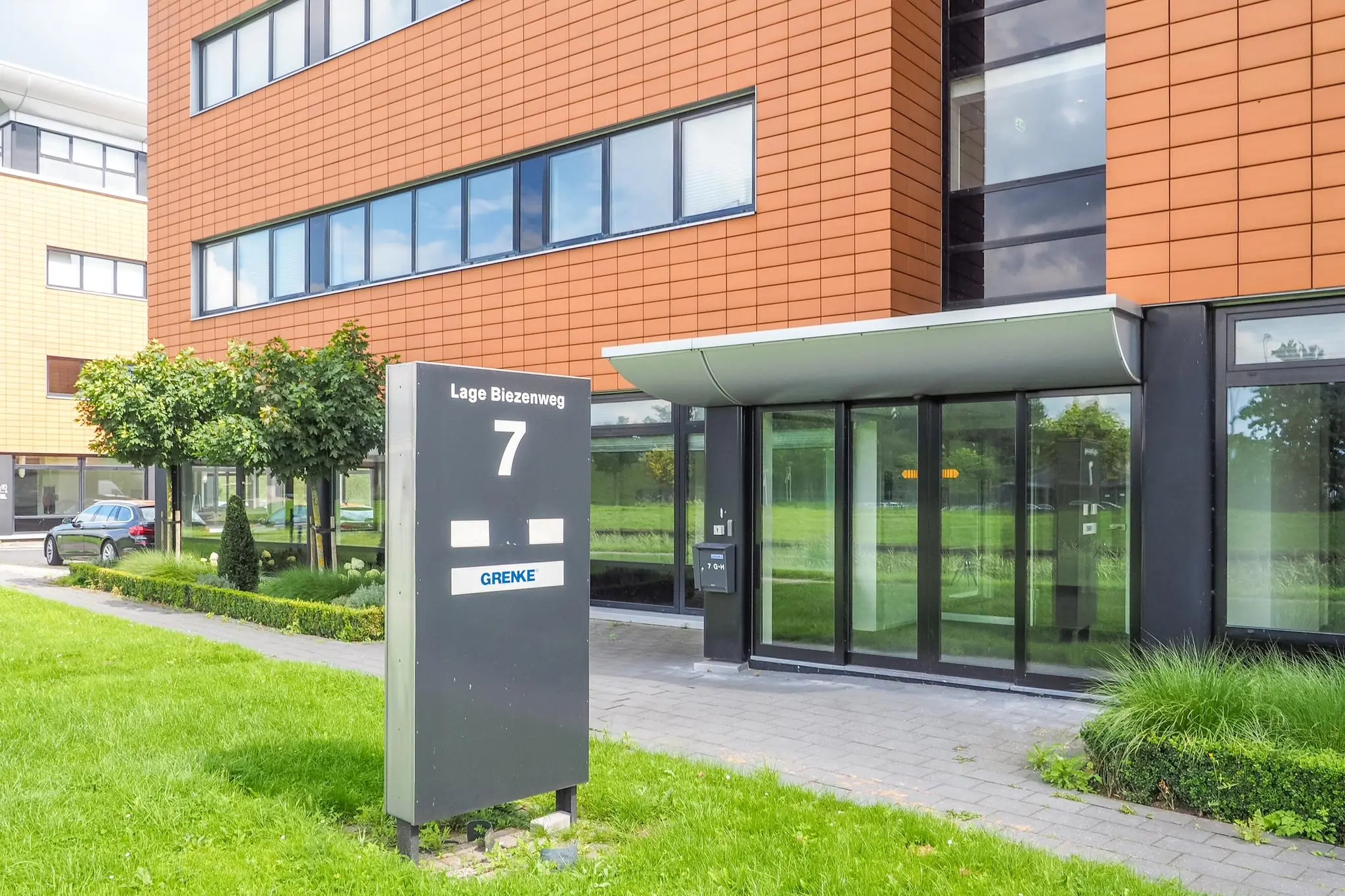 Ingang van een modern kantoorgebouw aan de Lage Biezenweg 7 met een bewegwijzeringsbord en groene omgeving.